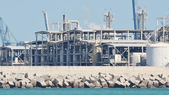 Smoky oil refinery complex, metal structures, and storage tanks on a pier with breakwaters. Marine terminal for receiving and distributing oil from tankers.