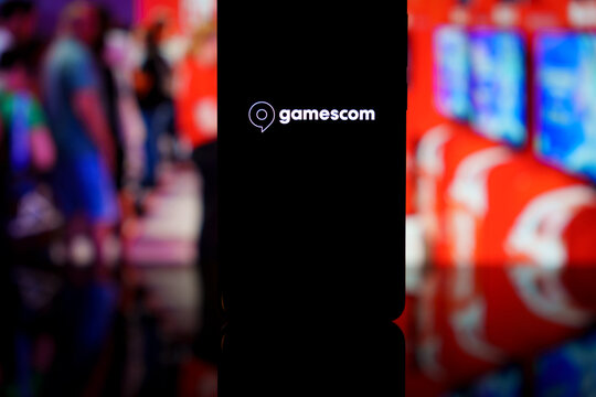 Gamescom logo displayed on a smartphone screen with a blurred gaming exhibition background, representing video game industry trade fairs and mobile coverage of gaming events