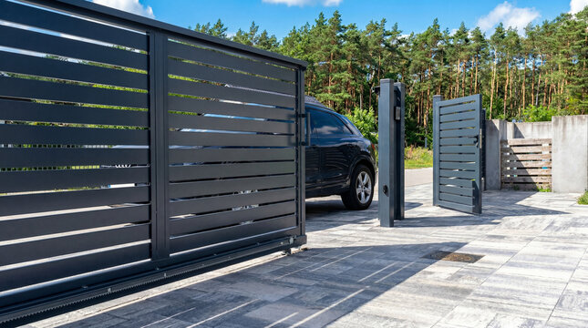 Modern dark gray horizontal slat automatic sliding gate system for a residential driveway. Features a sleek design, electronic keypad, and luxury car parked on stone pavers under a blue sky.