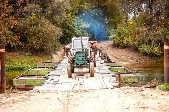 Ukraine : Small agricultural tractor paused without driver on old wooden bridge over river, smoke rising from exhaust in autumn village setting.