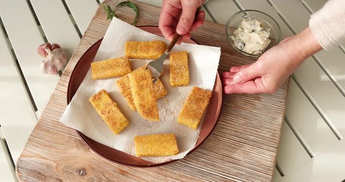 Crispy fried polenta sticks on plate with parchment paper. Hand using fork to pick golden cornmeal appetizer served on rustic wooden board with garlic and dipping sauce.