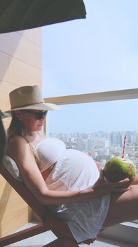 Expectant mother enjoying a sunny day on a city balcony while drinking fresh coconut water
