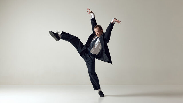 Dancer performing high kick jump in formal suit isolated on neutral background. Concept of breaking routine, high energy performance, confident self expression and dynamic corporate lifestyle
