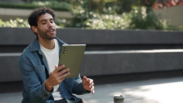 Portrait of curly haired Hispanic man talks on video call via digital tablet in city park. Latin guy discusses college news with friend on online chat via device as sits on parapet