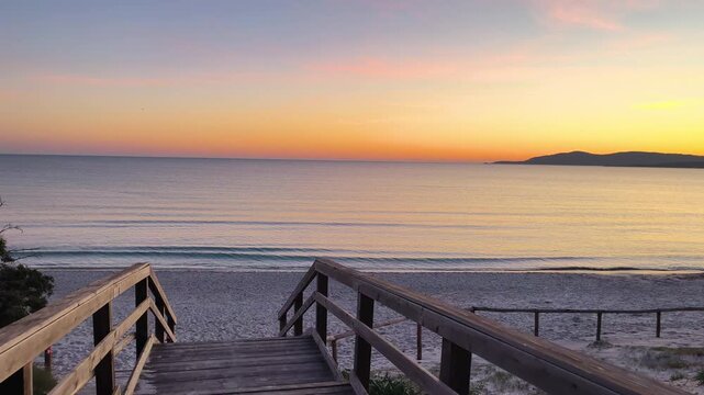 Slow motion point of view walk along a wooden boardwalk leading to the beach at sunset in Maria Pia, Alghero, Sardinia. Warm golden light, soft shadows and natural Mediterranean coastal atmosphere.
