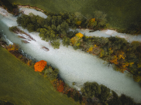 Aerial view of a serene river meanders through vibrant autumn foliage, a tapestry of greens and oranges against the muted water, Bohinjska Bistrica, Slovenia.