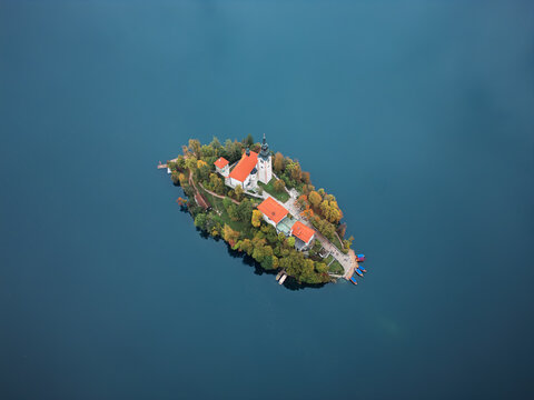Aerial view of the iconic island church with its red-tiled roofs, nestled amidst the sapphire waters of Lake Bled, surrounded by trees in autumn colors, Bled, Radovljica, Slovenia.