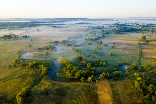 Aerial view of a serene Nida river meanders through a misty landscape, the morning sun casting a golden glow over the tranquil scene, Motkowice, Swietokrzyskie Voivodeship, Poland.