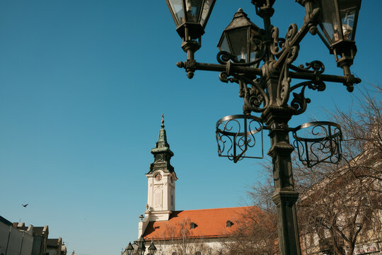 Historic church tower with decorative street lamp in foreground under blue sky in Sombor, Serbia, urban European architecture