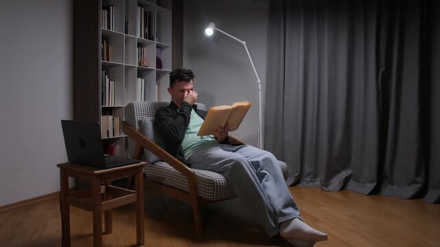 Young man rubbing tired eyes from eye strain while reading a book under a lamp at night in a living room, showing fatigue, studying, poor lighting and exhaustion