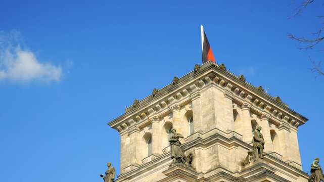 BERLIN, GERMANY - MARCH 23 2026: Static low angle shot of a historic tower of the Reichstag building featuring stone sculptures and the German national flag against a blue sky