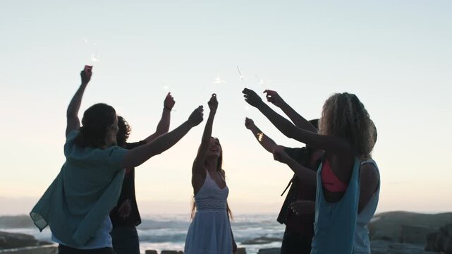 People, friends and dance at beach with sparklers in evening for goals, birthday or smile on vacation. Men, women and group with fireworks, excited and celebration on seaside getaway for milestone