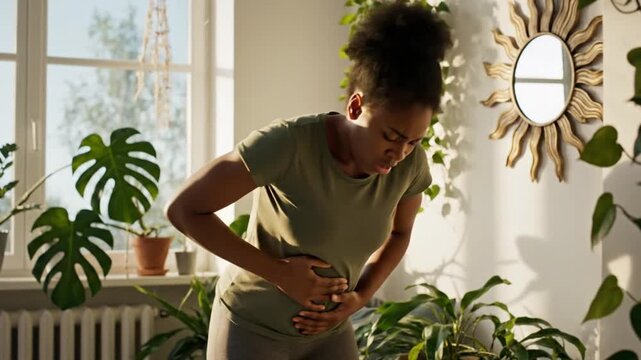 A young woman wearing a green t-shirt clutching her stomach in severe physical pain, expressing discomfort from abdominal cramps, digestive issues, or menstruation.