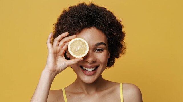 Happy young woman holding lemon slice over eye smiling on yellow background healthy lifestyle