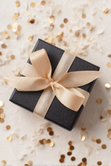 Fototapeta premium Elegant black gift box with gold satin ribbon surrounded by festive golden confetti on a textured cream background, perfect for celebrations or luxury occasions.