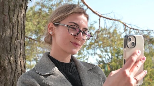 A young woman is outdoors, interacting with her smartphone as she adjusts her hair and poses for the camera