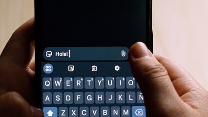 Close-up of hands holding a smartphone while typing "Hola!" (Hello!) in a messaging app. © Baudalab