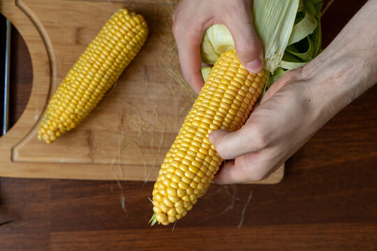 Male hands holding and shucking  fresh corn on the cob on a wooden table.