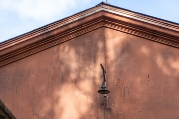 Old metal outdoor lamp hangs on triangular facade of brown plastered building featuring tree shadows weathered texture architectural cornice blue sky background. © Dmitry