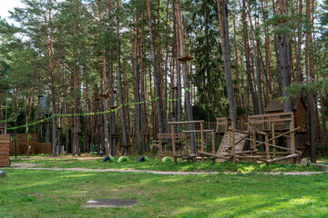 Obraz premium High ropes adventure park nestled in coniferous forest features wooden climbing structures platforms suspended bridges safety cables green bunting flags grassy lawn tall pine trees sunny daylight.