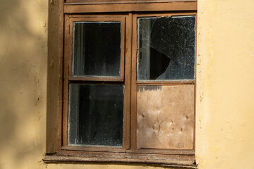 Obraz premium Weathered wooden window frame set into yellow peeling plaster wall featuring shattered glass shards and plywood board repair in derelict building structure.