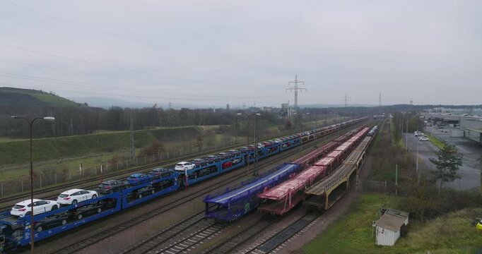Wide aerial view of a long freight train transporting new cars on specialized auto rack wagons through an industrial area
