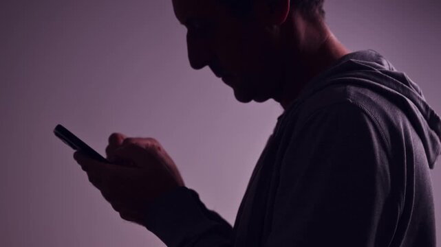 Mature man wearing hoodie using smartphone in low key light, concept of privacy, anonymity, or online communication. 4K.