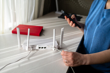 Obraz premium Woman Using Smartphone to Configure White WiFi Router While Connecting Ethernet Cable at Home