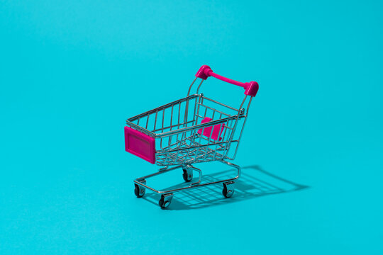 Minimalist photo of deep pink push cart as concept of supermarket, retail and sale season. Shopping trolley on turquoise blue background central composition.