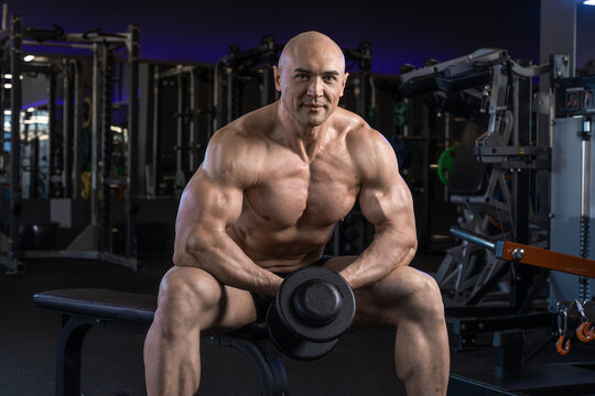 Muscular man holds a dumbbell while seated on a bench in a gym.