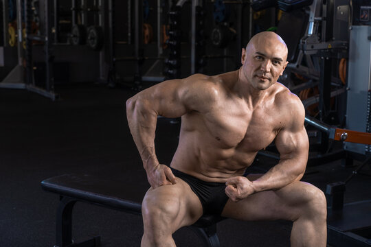Muscular man poses confidently on a bench within a gym setting.