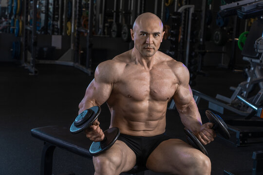 Muscular man sits on a bench in a gym, lifting dumbbells and showcasing his physique.