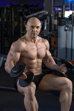Muscular man sits holding dumbbells in a gym, showcasing strength and fitness.