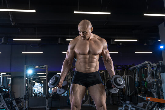 Muscular man holds dumbbells while posing in a gym with mirrors and equipment.