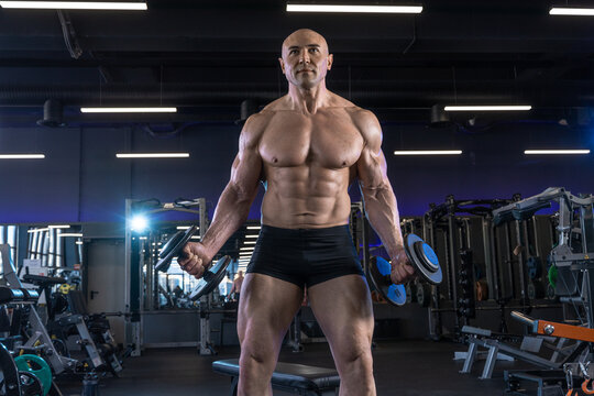 Muscular man stands confidently in a gym, holding dumbbells and showcasing his physique.