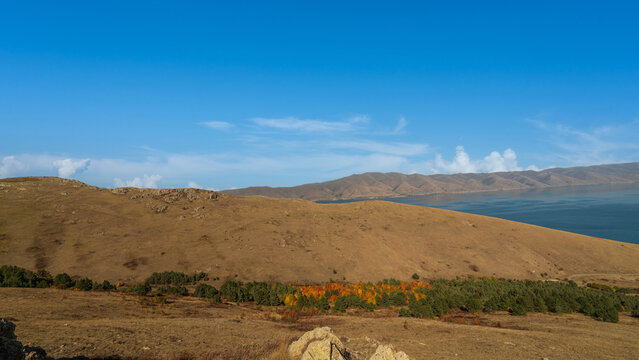 Mountain slope above Lake Sevan in Armenia during autumn. Golden fields and trees with orange foliage contrast against the deep blue water and sky.