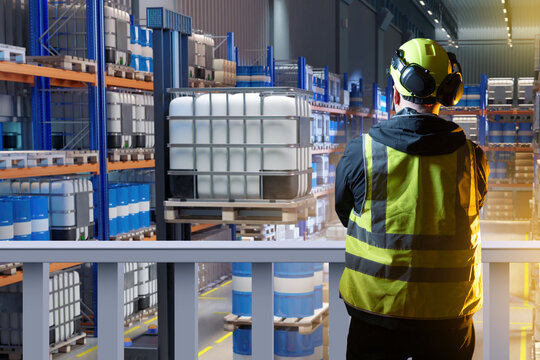 Industrial warehouse storage of chemicals. Worker in safety gear oversees stacked drums and IBC containers on shelving, representing hazardous materials management, logistics, and inventory control.