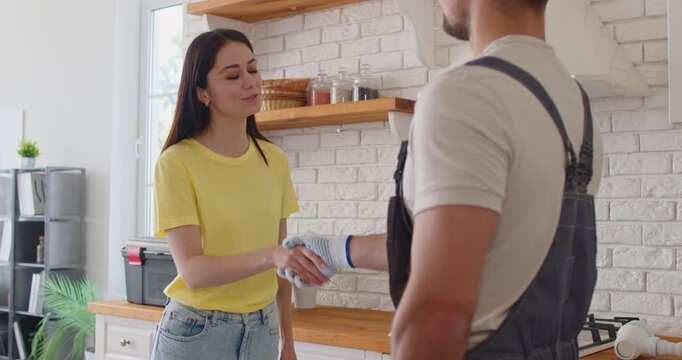 Professional repairman in uniform shaking hands with woman after successful repair standing in kitchen. Man plumber with client. Plumbing maintenance concept. 4k video. Slow motion video.