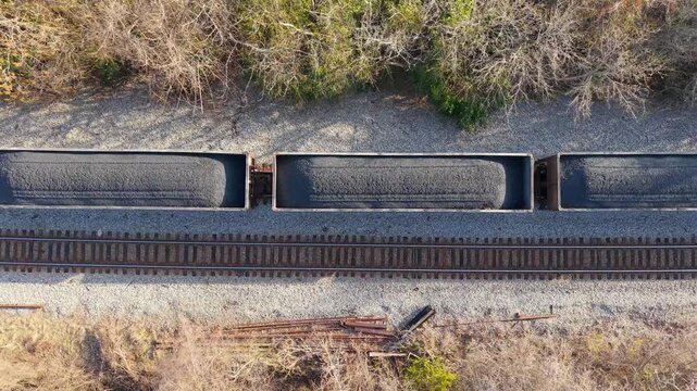 Aerial top-down video of a freight train transporting open-top hopper cars filled with coal along a rural railway line.