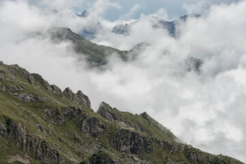 Fototapeta premium A dramatic view of rugged mountain ridges and peaks partially shrouded in heavy fog and clouds under an overcast sky, showcasing high-altitude wilderness.