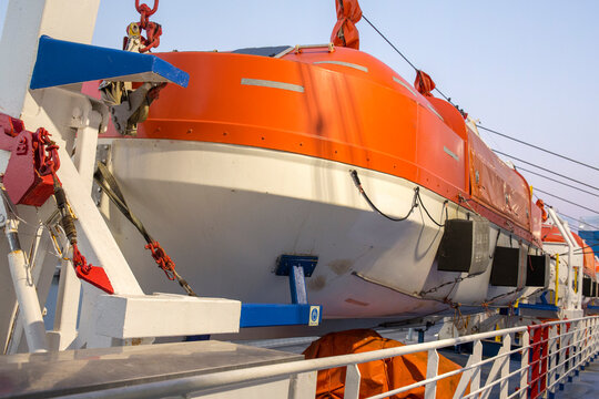 An orange and white lifeboat is mounted on a large ship's deck, secured by metal davits and rigging. The scene highlights essential maritime safety equipment used for emergencies on the open sea.