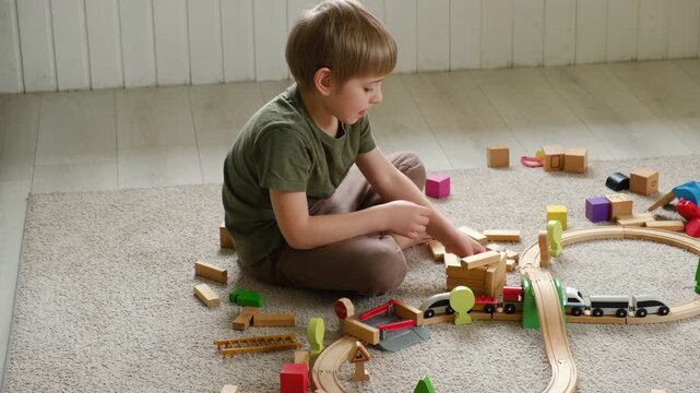 Cute little blonde child boy sitting on the floor playing with wooden toy train. Toddler kid plays railway. Made of natural materials. Birthday gift. Preschool employment. Brain, coordination exercise