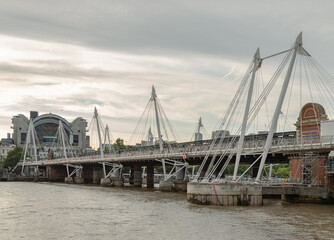 A striking architectural dialogue unfolds across the River Thames, where the industrial legacy of the Hungerford Bridge meets the modern elegance of the Golden Jubilee Footbridges. The central railway