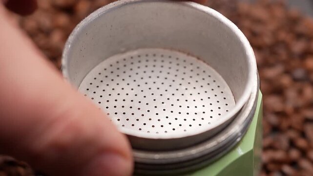 Putting aluminium sieve into moka pot coffee maker over coffee beans background
