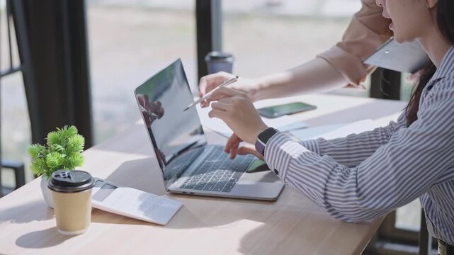 Close-up of two businesswomen working on a laptop, one pointing at the screen while the other types, showing teamwork, guidance, and collaboration in a bright office.