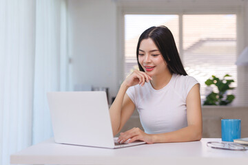 Fototapeta premium Young happy asian woman smiling and using a laptop computer while studying or working remotely from her home office desk