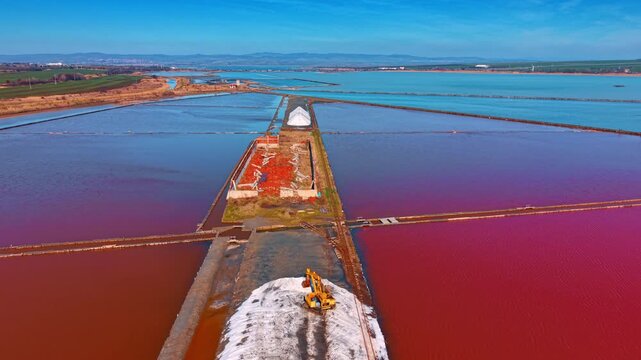 Aerial view of pink salt evaporation ponds with industrial ruins. Wide perspective of colorful salt pans, white salt piles, and an abandoned brick structure under blue sky.