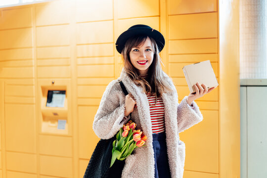 Portrait of smiling fashion woman with box near modern postal automatic mail terminal with self service device for pickup or refund an order. Electronic locker for storing parcels. Selective focus