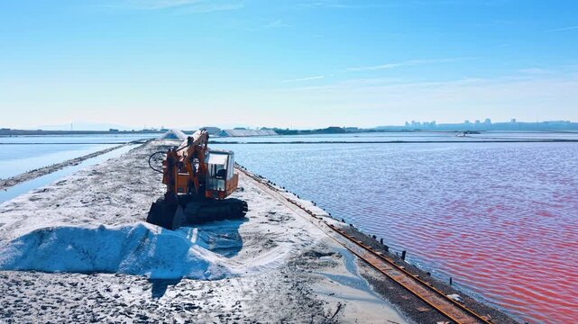 Yellow excavator on salt pile at pink salt lake evaporation pond. Construction machinery working on a dike between vibrant pink water basins under a clear blue sky.