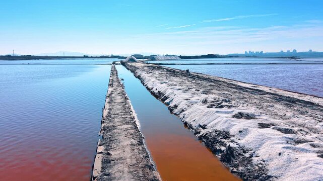 Perspective view of salt piles and dikes in pink water lagoon. Symmetrical lines of white salt heaps on a narrow path separating colorful evaporation ponds.